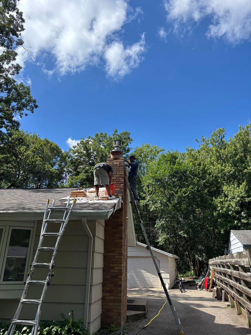 Two chimney technicians standing on ladders and the roof while rebuilding a brick chimney on a home in Saint Paul, Minneapolis MN. The workers are removing damaged bricks and repairing the upper section of the chimney under a clear blue sky, surrounded by trees.